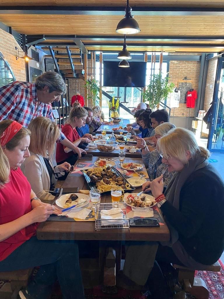 A group of tourists sitting and eating and a man pouring beer in glasses from the bottle at Pineios Brewery tasting room